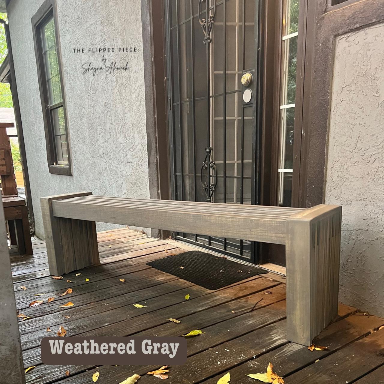 A wooden bench in weathered gray finish placed on a wooden porch. It's positioned near a dark metal door with a mesh screen. Fallen leaves are scattered on the porch. A logo in the corner reads "The Flipped Piece, Shayna Alnwick."