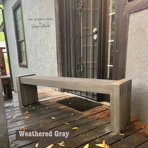 A wooden bench in weathered gray finish placed on a wooden porch. It's positioned near a dark metal door with a mesh screen. Fallen leaves are scattered on the porch. A logo in the corner reads "The Flipped Piece, Shayna Alnwick."