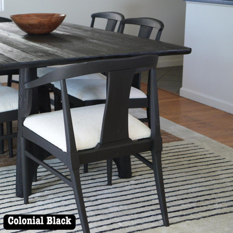 Black wooden dining table with matching chairs featuring white cushions, set on a striped rug. A wooden bowl sits on the table. The room has light wooden floors and a partial view of a kitchen in the background. Label reads "Colonial Black."