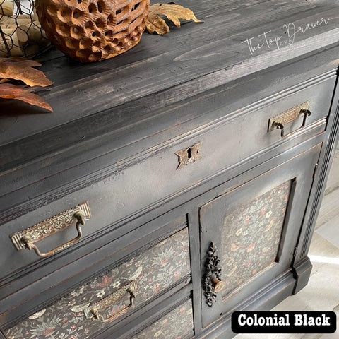 A black wooden dresser with ornate drawer handles and floral patterned drawer fronts. A decorative bowl and autumn leaves rest on top. The words "The Top Drawer Rva" are visible, and "Colonial Black" appears at the bottom.