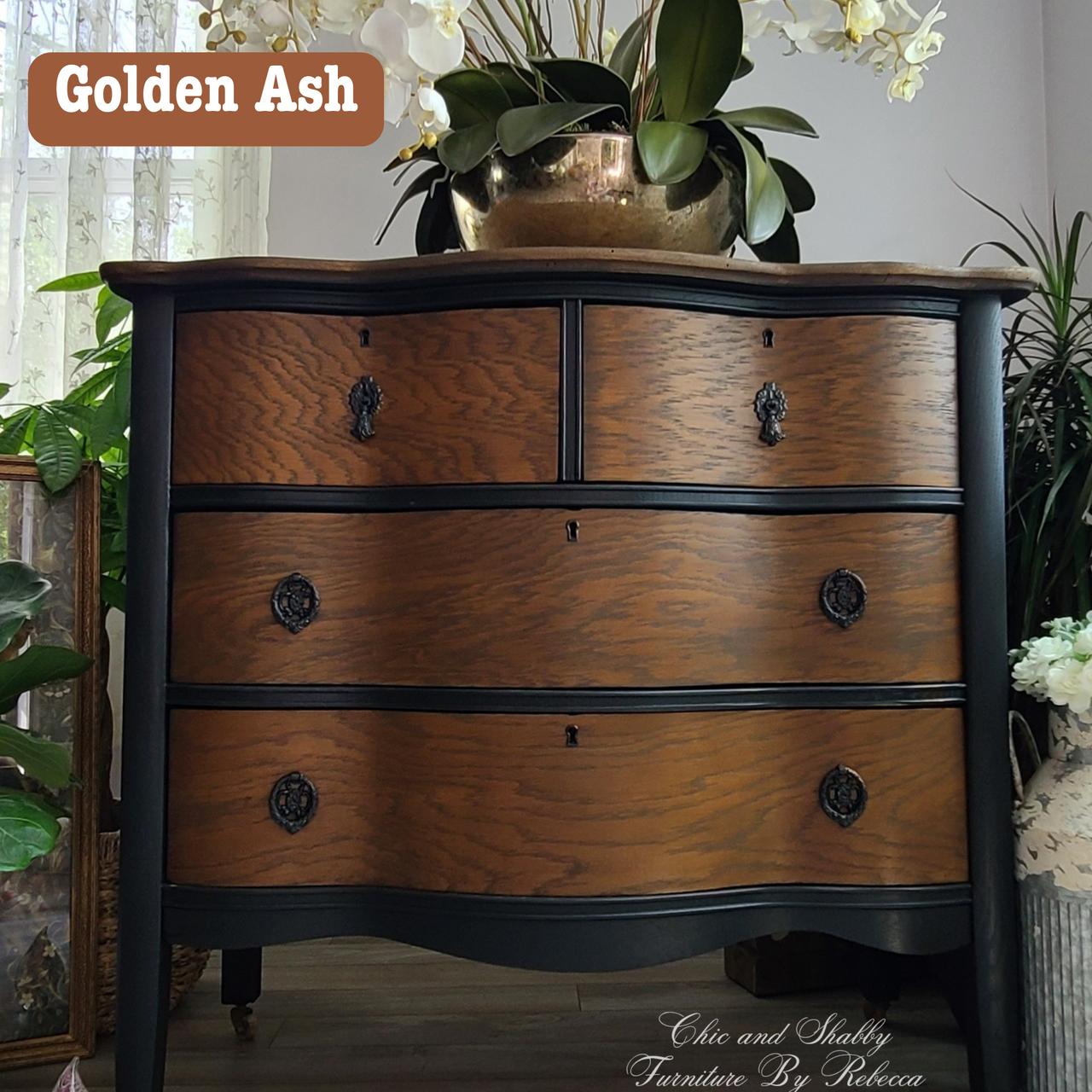 A vintage wooden dresser with four drawers, featuring dark wood accents and elegant metal handles. A vase with orchids and other plants surround it. A label reads "Golden Ash" at the top left corner.