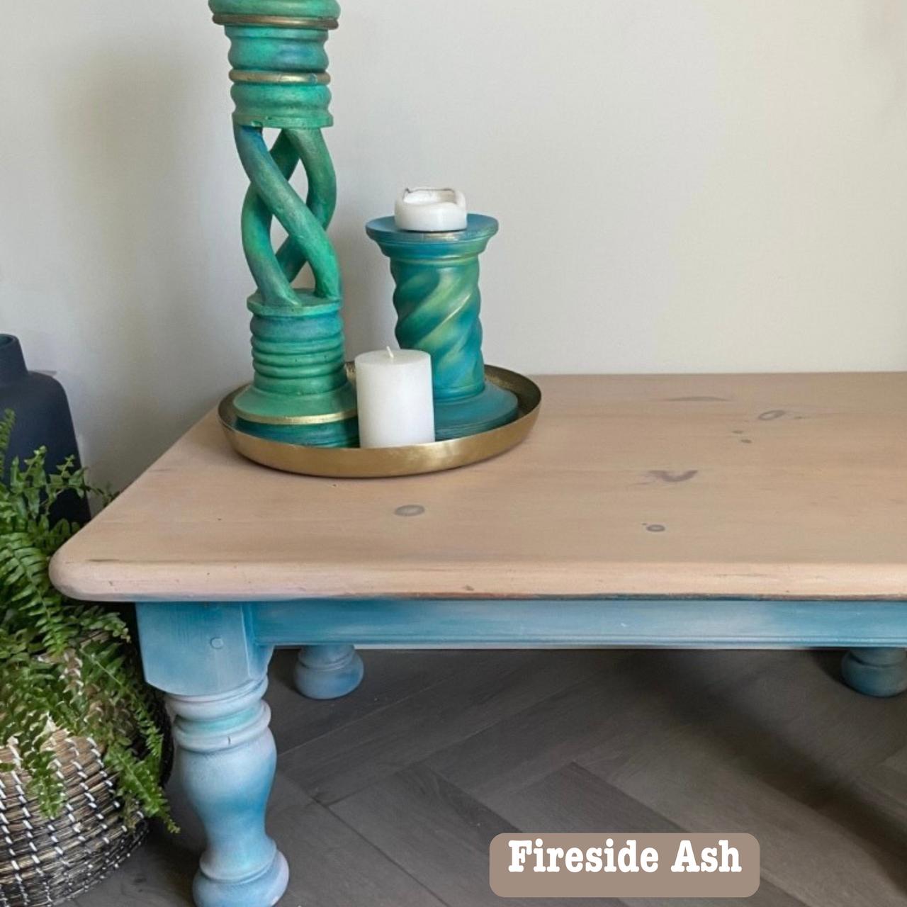 A wooden table with blue legs holds a decorative arrangement of tall, twisted teal candle holders, each with white candles on a brass tray. Nearby, there's a plant in a woven basket. A simple, modern interior design setup.