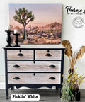 A rustic dresser with a desert landscape painting, featuring Joshua trees, above it. The dresser has black accents and a natural wood finish. Decorative items include candles, a vase with yellow flowers, and branding text for Theresa Lee NTS Design Co.