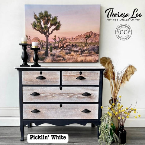 A rustic dresser with a desert landscape painting, featuring Joshua trees, above it. The dresser has black accents and a natural wood finish. Decorative items include candles, a vase with yellow flowers, and branding text for Theresa Lee NTS Design Co.