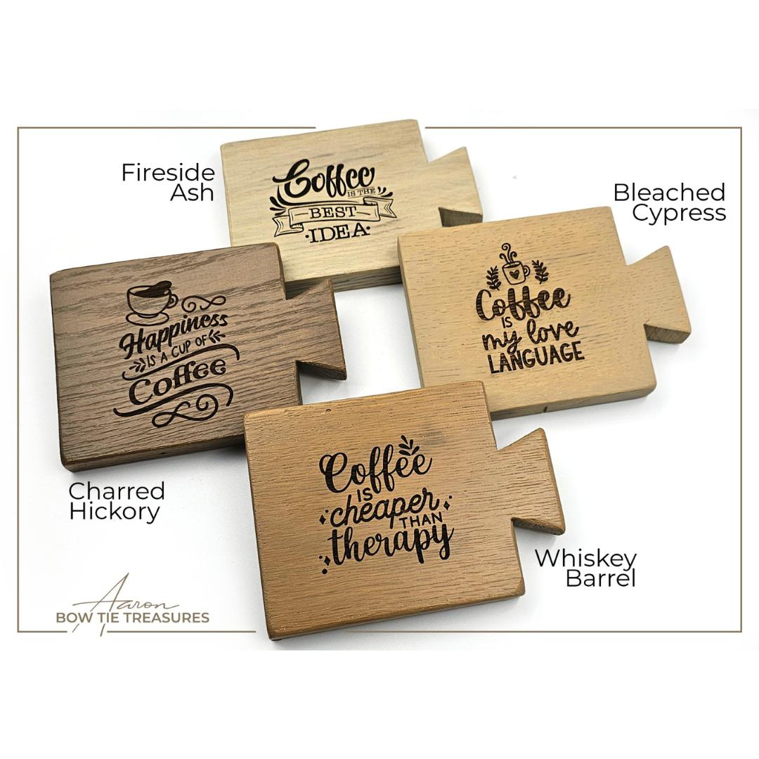 Four wooden coasters with coffee-themed engravings. Designs include "Happiness is a cup of coffee," "Coffee is my love language," "Coffee is cheaper than therapy," and "Coffee is the best idea." Coasters are made from various wood types.
