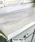A wooden cabinet countertop featuring a distressed, pickling white finish. The surface has a weathered look, with visible brush strokes. A green plant leaf is partially visible in the foreground.