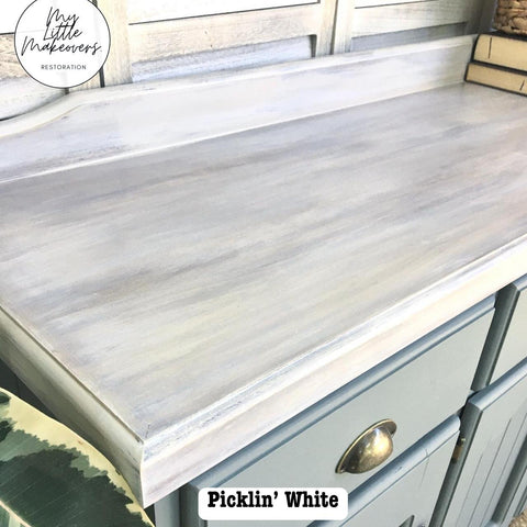 A wooden cabinet countertop featuring a distressed, pickling white finish. The surface has a weathered look, with visible brush strokes. A green plant leaf is partially visible in the foreground.