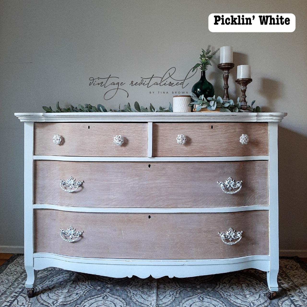 Vintage wooden dresser with a mix of natural wood and distressed white paint. It has decorative white knobs and drawer handles. The top is adorned with green leaves, a green glass bottle, and two white candlesticks. Rug underneath.