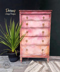 A dresser that has multiple colors of chalk paint from flamingo to peony to fluff all blended beautifully.  there is a plant to the left in a black pot and a rug in front