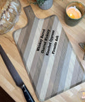 A wooden cutting board with diagonal stripe patterns and labeled wood types: Whiskey Barrel, Charred Hickory, Bleached Cypress, and Fireside Ash. It is on a countertop with a serrated knife, a loaf of bread, a lit candle, and greenery nearby.