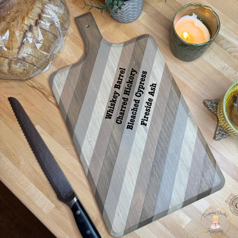 A wooden cutting board with diagonal stripe patterns and labeled wood types: Whiskey Barrel, Charred Hickory, Bleached Cypress, and Fireside Ash. It is on a countertop with a serrated knife, a loaf of bread, a lit candle, and greenery nearby.