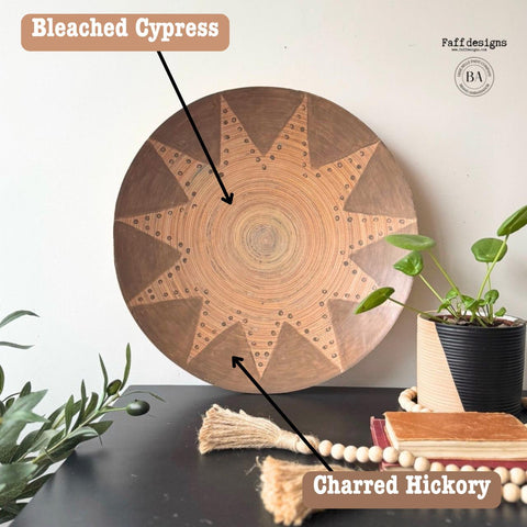 A decorative wooden bowl with a sunburst pattern is displayed on a dark surface. Nearby, a potted plant, decorative beads, a stack of books, and a tassel add to the arrangement. The wall is plain, highlighting the bowl's intricate design.