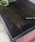 A wooden surface stained in a dark shade labeled "Colonial Black," showcasing a smooth, glossy finish with visible wood grain. A plant and scattered pages are partially visible in the background.