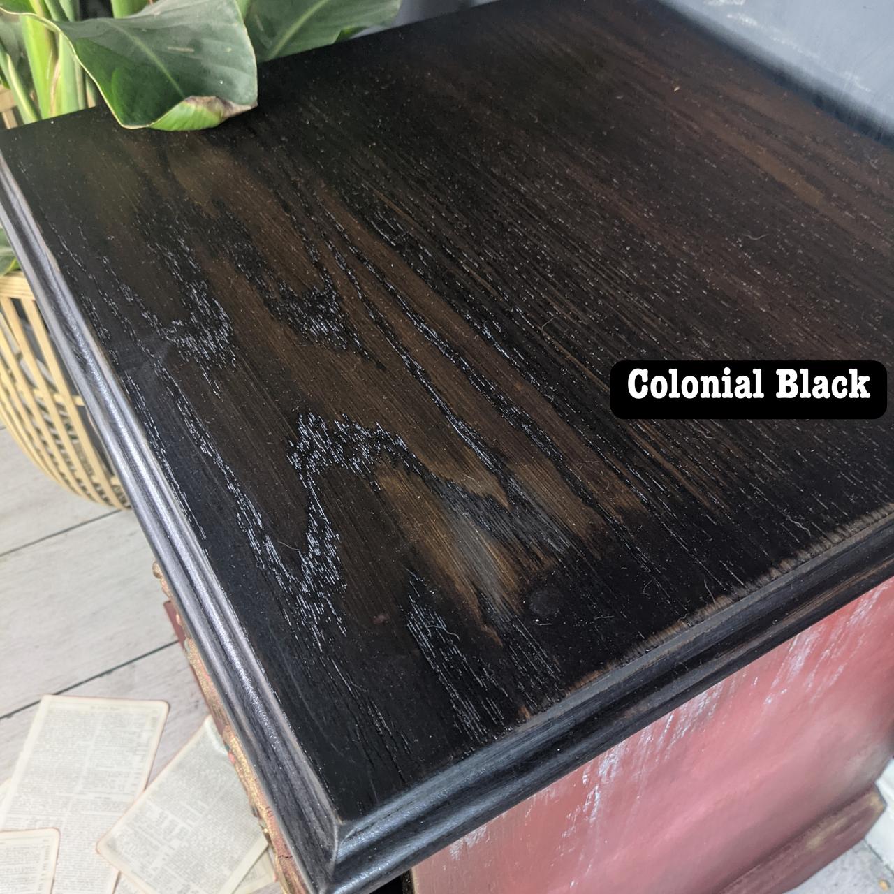 A wooden surface stained in a dark shade labeled "Colonial Black," showcasing a smooth, glossy finish with visible wood grain. A plant and scattered pages are partially visible in the background.