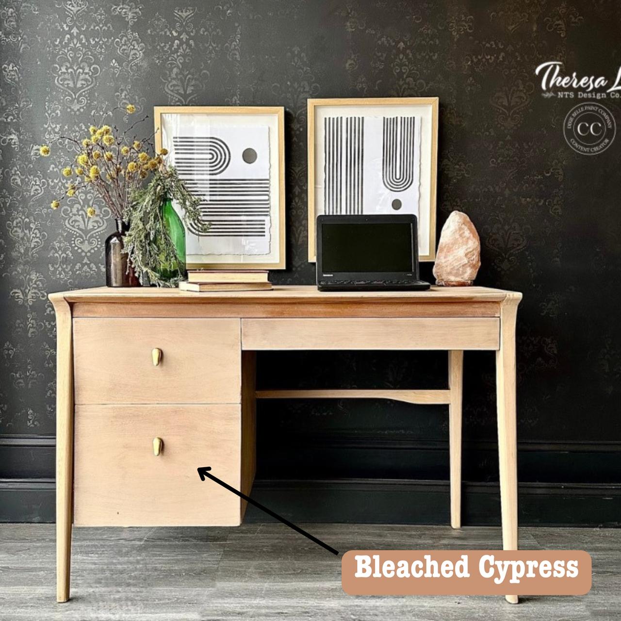 A wooden desk with two drawers stands against a dark patterned wall. It holds a laptop, a salt lamp, yellow flowers in a glass vase, and two framed abstract artworks. The floor is grayish and textured.