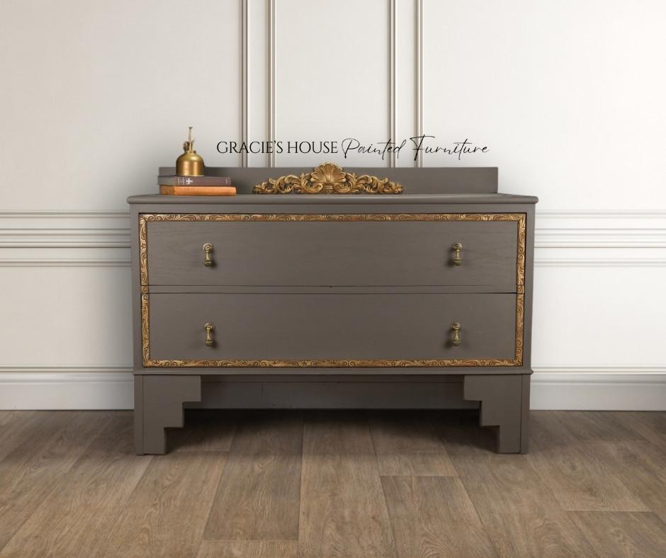 Dixie Belle Paint Gravel Road Chalk Mineral Paint is the perfect paint for any DIY project! Gray dresser with gold trim and ornate handles, set against a white paneled wall. A decorative gold ornament rests on top, along with a small gold bottle and a framed sign reading "Gracie's House Painted Furniture." Elegant wood floor below.