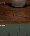 Close-up of a wooden dresser top with a rich, dark espresso finish and subtle texture. The top shows natural wood grain, contrasting with a deep green-painted front panel below. A round ceramic pot is partially visible on the top.
