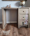 A vintage wooden desk with three drawers adorned with painted floral designs. A small globe and two decorative deer figurines sit on top. A textured wall and framed art are in the background, with logos in the bottom left corner.