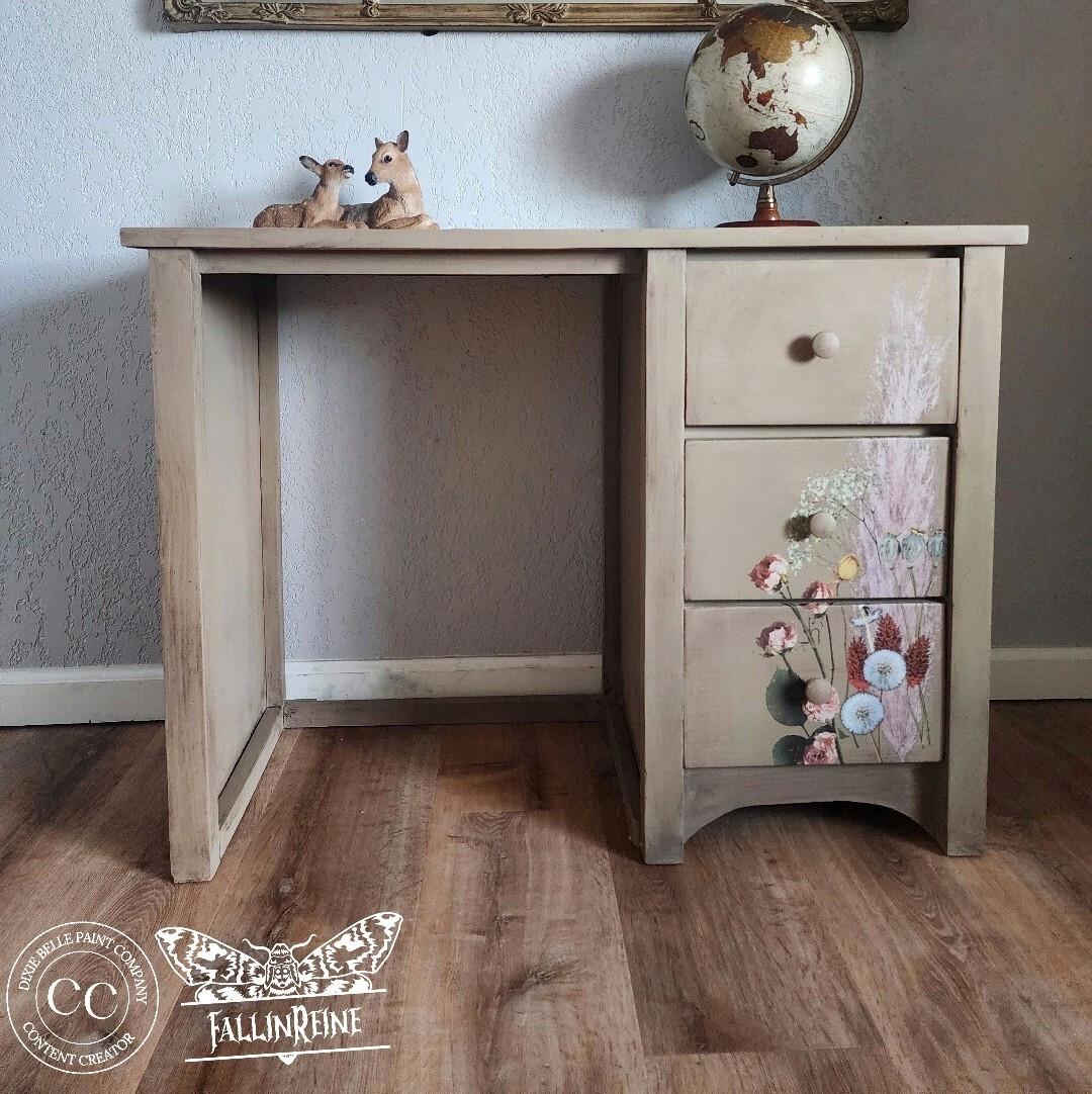 A vintage wooden desk with three drawers adorned with painted floral designs. A small globe and two decorative deer figurines sit on top. A textured wall and framed art are in the background, with logos in the bottom left corner.