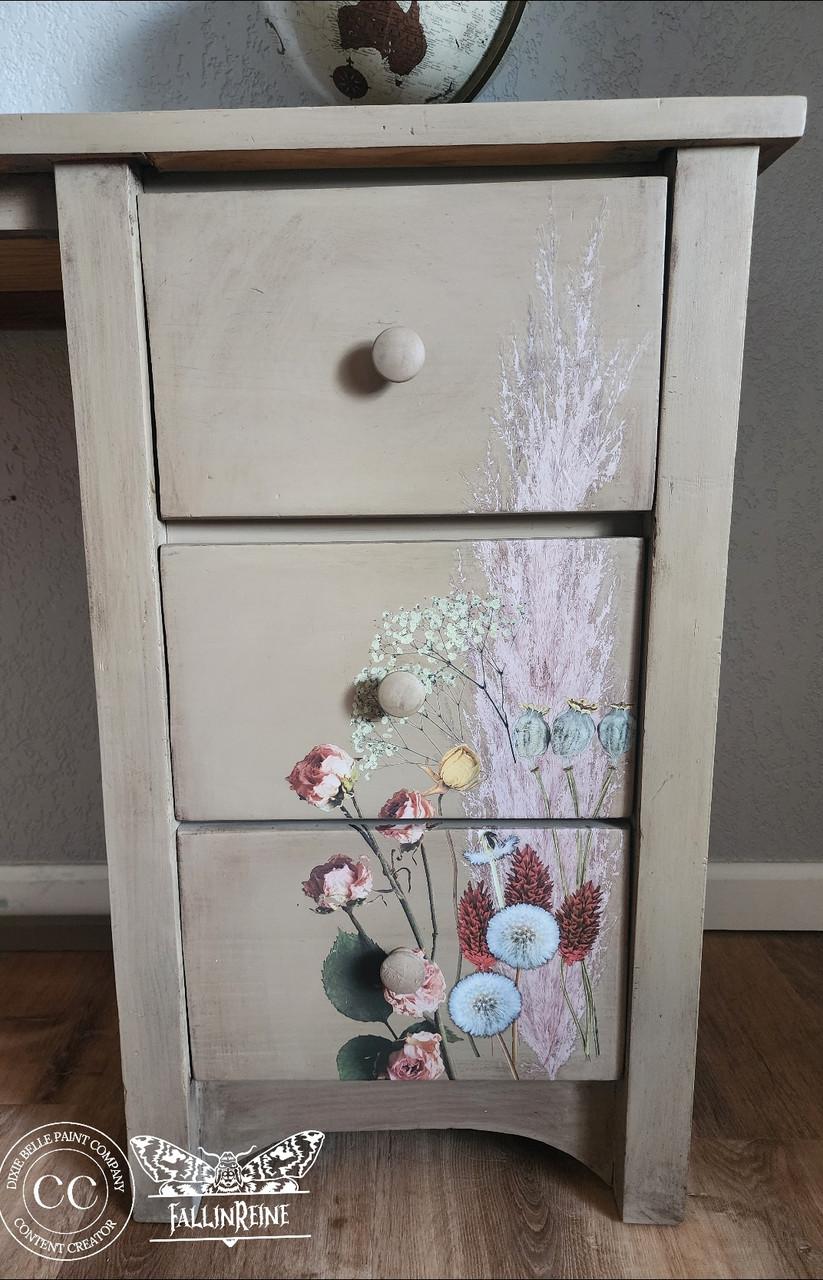A wooden dresser with three drawers. The front showcases a floral design with various flowers in soft colors. A logo featuring the text "FallinReine" and a circular emblem are on the bottom left. The dresser stands on a wooden floor.