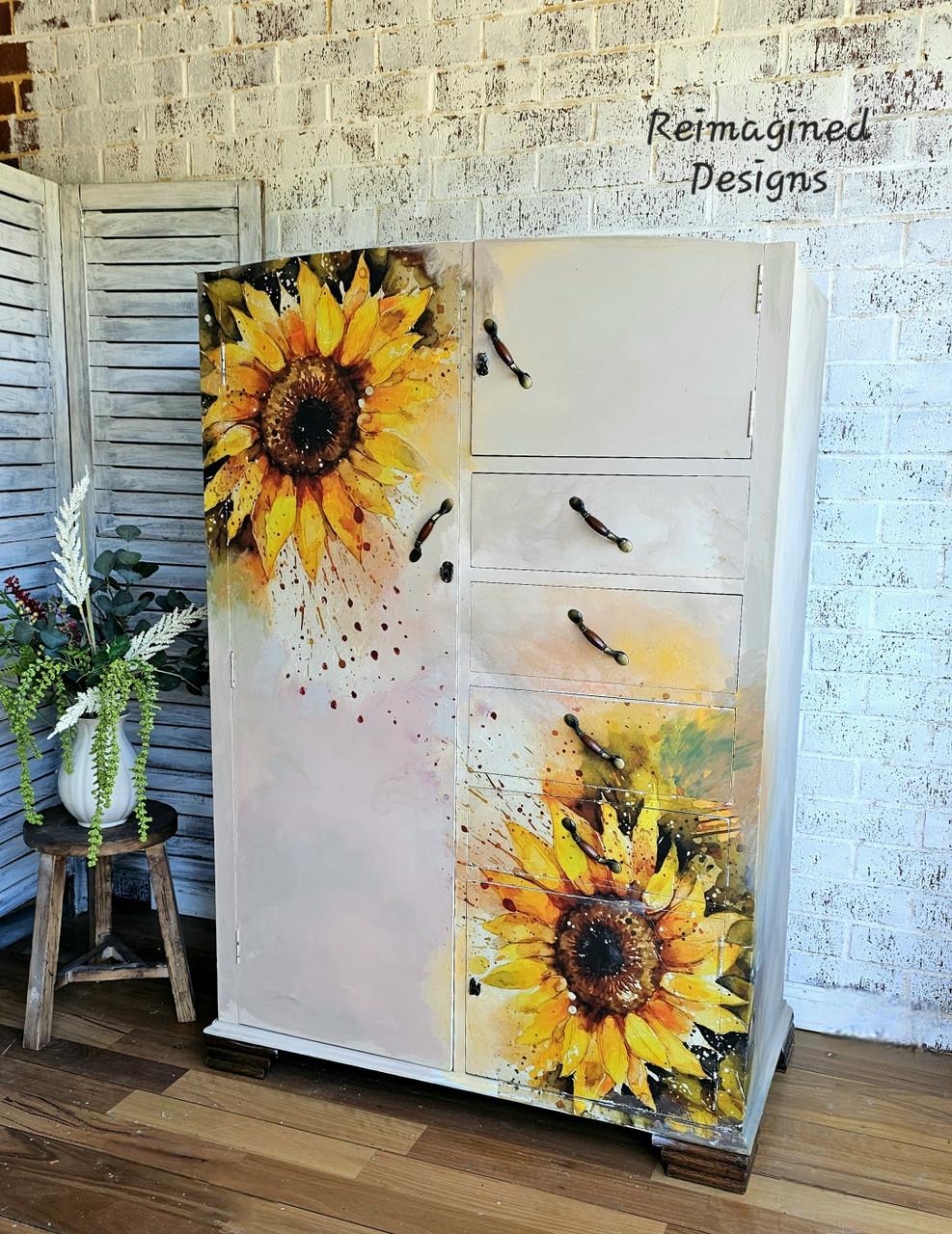 White furniture piece with large, vibrant sunflowers. It features several drawers and a cabinet, set against a white brick wall. A small potted plant is placed nearby on a wooden stool and a folding screen is visible on the left.