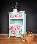 A small vintage cabinet painted in light blue with floral designs on the drawer and door. It stands on a wooden floor against a dark background. A white jug with flowers sits on top, and a wicker basket with flowers is beside it.
