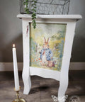 White wooden nightstand with a decoupage of a rabbit in a blue jacket on the front panel. A lit candle in a holder stands nearby. The scene has a whimsical, garden setting, and a small plant is on the tabletop. A watermark is visible at the bottom.