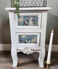 A white vintage-style nightstand with two drawers featuring floral and rabbit patterns. It has decorative legs and a detailed floral design at the bottom. A lit white candle in a gold holder stands beside it on a wooden floor.