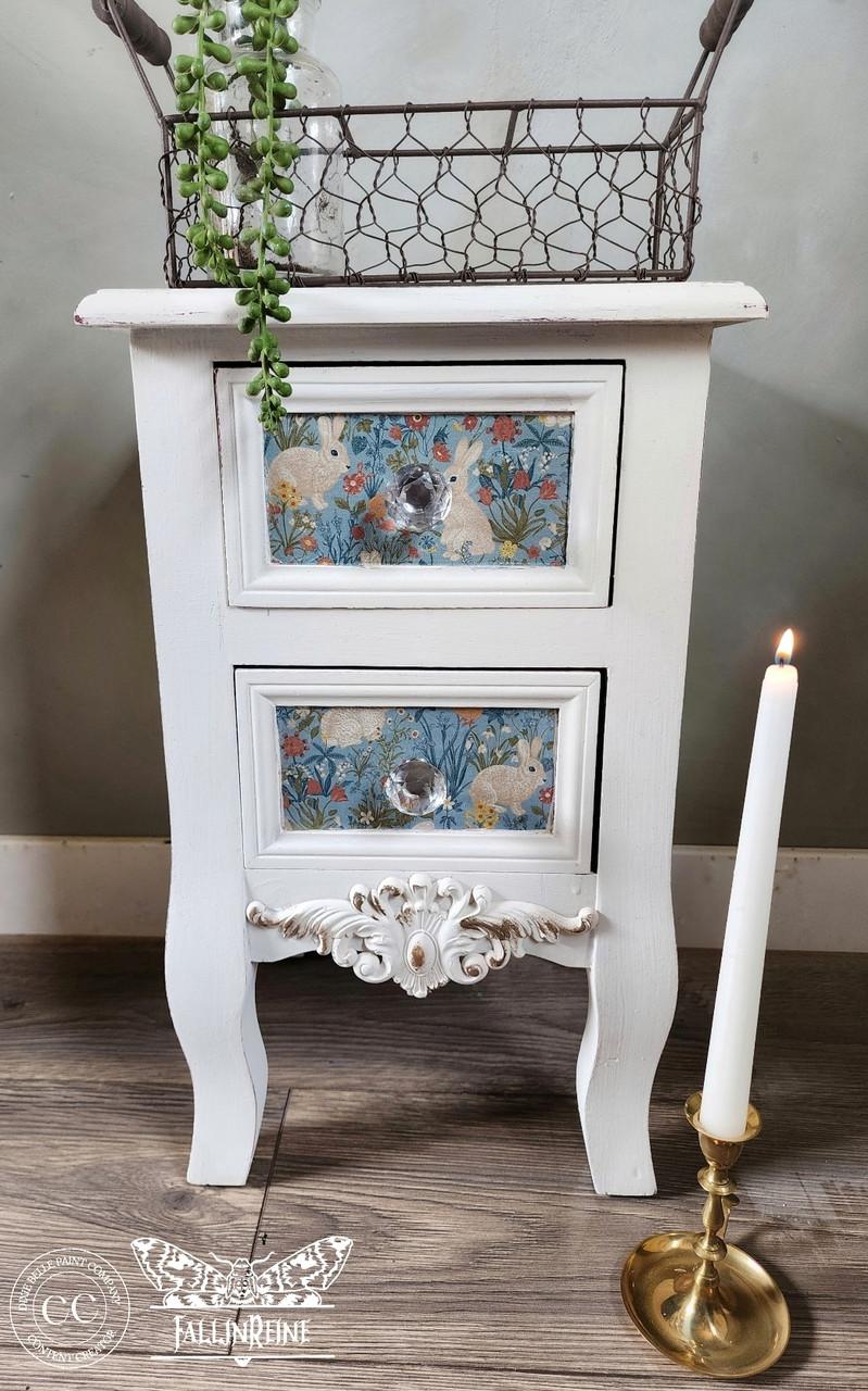 A white vintage-style nightstand with two drawers featuring floral and rabbit patterns. It has decorative legs and a detailed floral design at the bottom. A lit white candle in a gold holder stands beside it on a wooden floor.