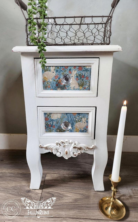 A white vintage-style nightstand with two drawers featuring floral and rabbit patterns. It has decorative legs and a detailed floral design at the bottom. A lit white candle in a gold holder stands beside it on a wooden floor.