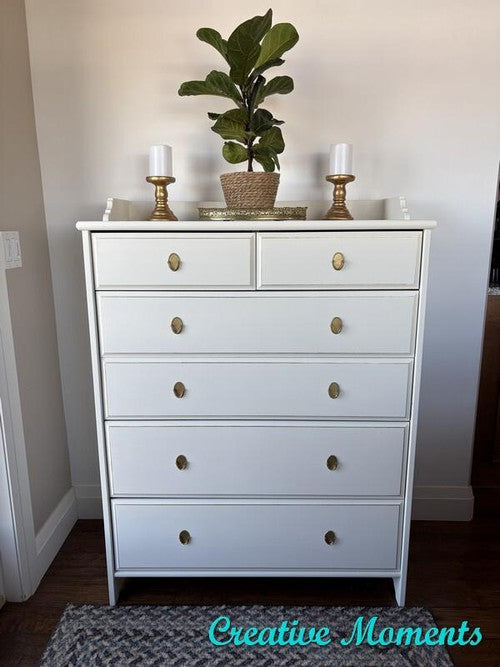 White dresser with six drawers and gold knobs, topped with a potted plant and two white candles in gold holders, standing against a light-colored wall with "Creative Moments" text in the bottom right corner.