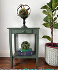 A small green side table with a single drawer displays a metal and moss globe on top. A potted fiddle leaf fig sits beside it, while a decorative book and faux plant rest on the table’s lower shelf. "Creative Moments" is written in turquoise.