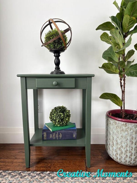 A small green side table with a single drawer displays a metal and moss globe on top. A potted fiddle leaf fig sits beside it, while a decorative book and faux plant rest on the table’s lower shelf. "Creative Moments" is written in turquoise.