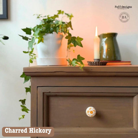 A brown wooden dresser with an ornate knob. A potted ivy plant, a lit candle, ceramic jug, and books are on top. Soft, warm lighting illuminates the scene. A logo for "faff designs" is in the upper right corner.