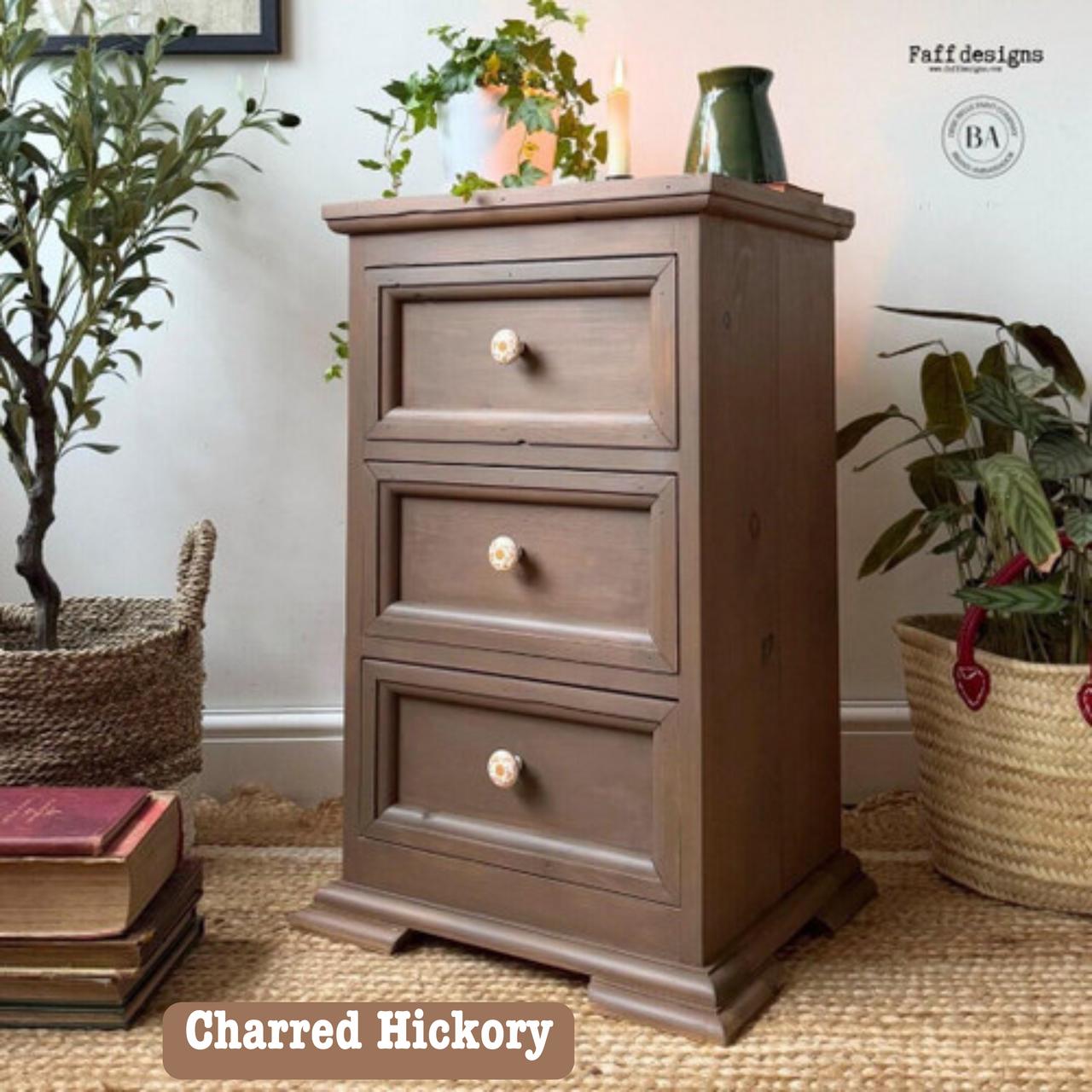 A wooden chest of drawers with three ornate knobs stands on a woven mat. It's surrounded by a potted plant, candles on top, a book, and baskets with plants inside. A small vase and a stack of books are placed nearby.