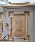 Antique wooden cabinet with ornate carvings on the door and a gold ring handle. The cabinet has a distressed gray and brown finish, with a curved top and a decorative mirror. It stands against a white brick wall.