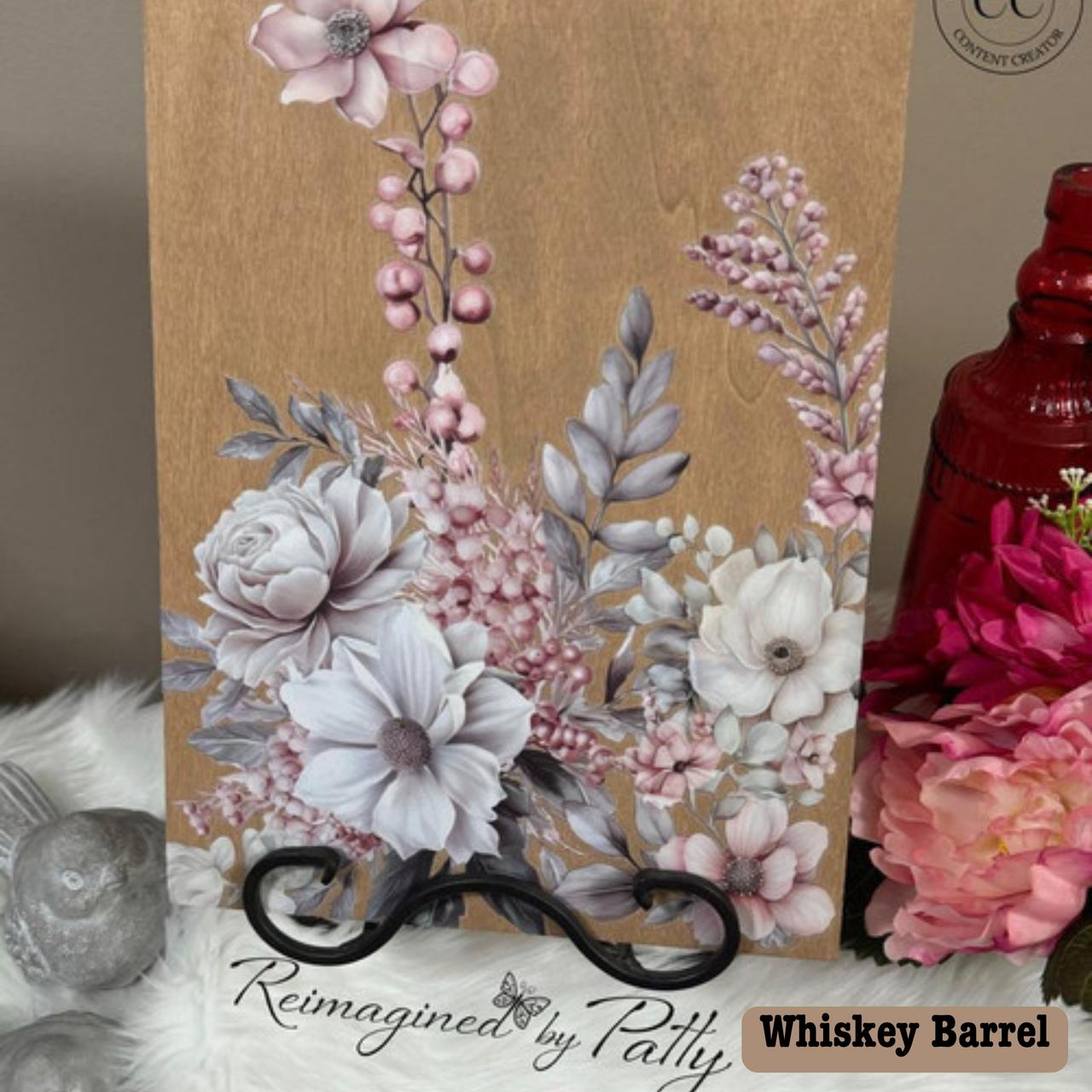 A wooden plaque with delicate floral artwork featuring pink and white flowers, with white leaves and small berries. It stands on a black holder, placed on a soft white surface. Text reads "Reimagined by Patty" in elegant script.
