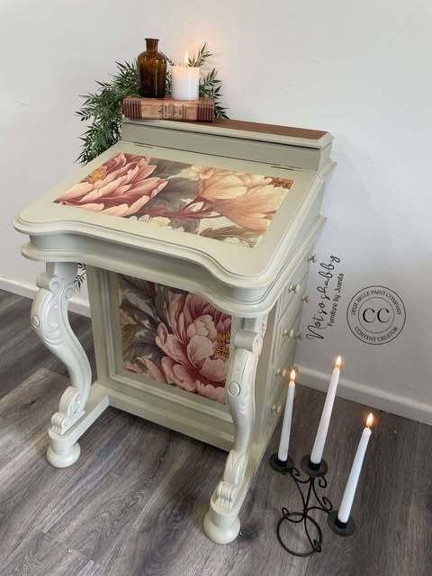 Vintage-style desk with floral patterns on the top and side. Books and lit candles are on the desk, with a decorative metal candle holder on the floor. The desk is set against a white wall on a wooden floor.