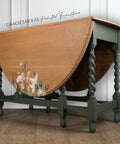 A wooden drop-leaf table with intricately carved green legs. One side has a floral design. The words "Gracie's House Painted Furniture" are visible at the top. The table is placed on a wooden floor against a light wall.