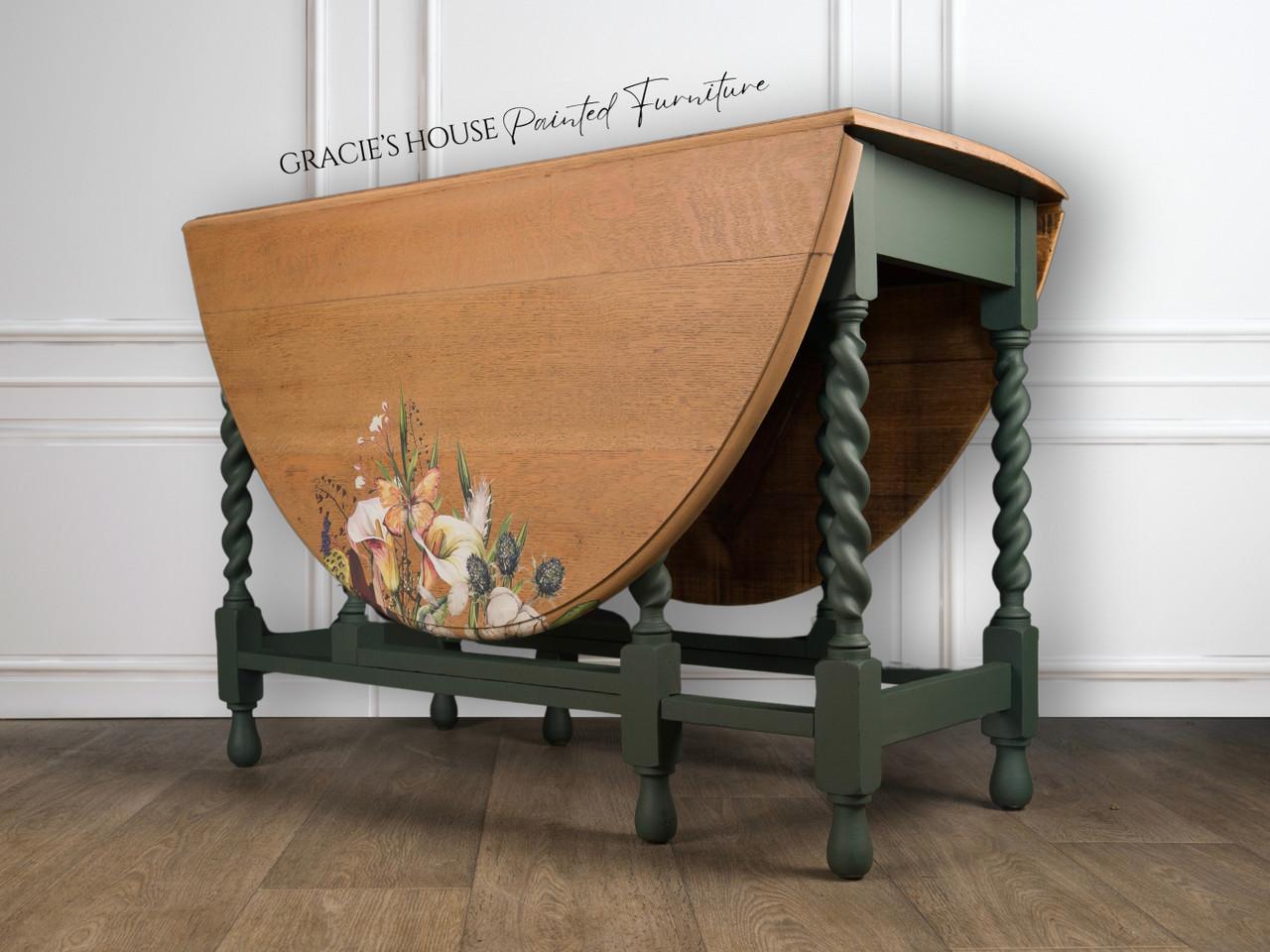 A wooden drop-leaf table with intricately carved green legs. One side has a floral design. The words "Gracie's House Painted Furniture" are visible at the top. The table is placed on a wooden floor against a light wall.