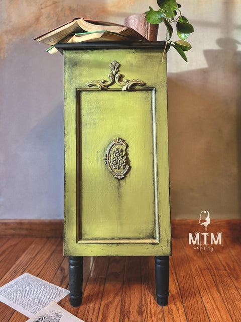 A small, ornate green cabinet with black wax accents stands on a wooden floor. An open book and a plant adorn the top. A paper is on the floor nearby.