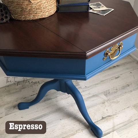 A hexagonal wooden table with a dark brown top and blue body, featuring a drawer with a brass handle. It stands on curved legs. The floor is distressed white wood. A wicker basket and books are partially visible on the table.