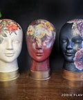 Three mannequin heads are painted with intricate floral designs and displayed on a wooden surface against a dark background. The left head is white with red and orange flowers, the middle is red with assorted flowers, and the right is blue with pink flowers. On the bottom right corner, the watermark "Jodie Flavell Art" is visible.