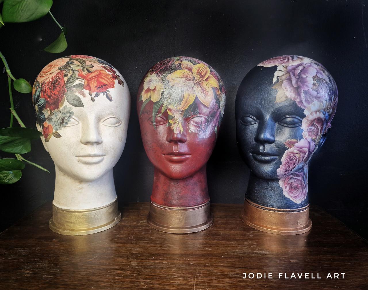 Three mannequin heads are painted with intricate floral designs and displayed on a wooden surface against a dark background. The left head is white with red and orange flowers, the middle is red with assorted flowers, and the right is blue with pink flowers. On the bottom right corner, the watermark "Jodie Flavell Art" is visible.