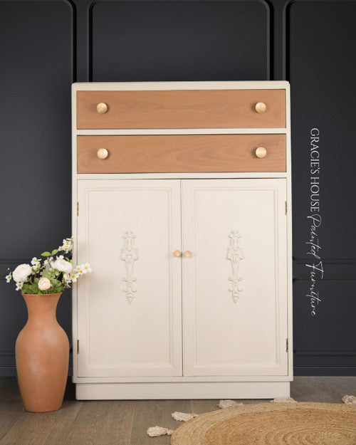 A cream-colored cabinet with ornate detailing and light wood drawers stands against a dark wall. Gold knobs accent the drawers and doors. A tan vase with white and yellow flowers sits next to the cabinet on a wood floor.