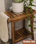 Wooden bathroom stand with a potted plant and draped towel on top, and another shelf below. The pot has a white textured design and the plant has trailing green vines. The text "Golden Ash" is in the corner.