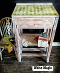 Vintage desk with a worn wood finish, featuring a green diamond pattern on top. A shelf holds an antique book and newspaper. A rustic chair sits beside it, adorned with leaves. A small, decorative cup is placed on the shelf. Text reads "White Magic."