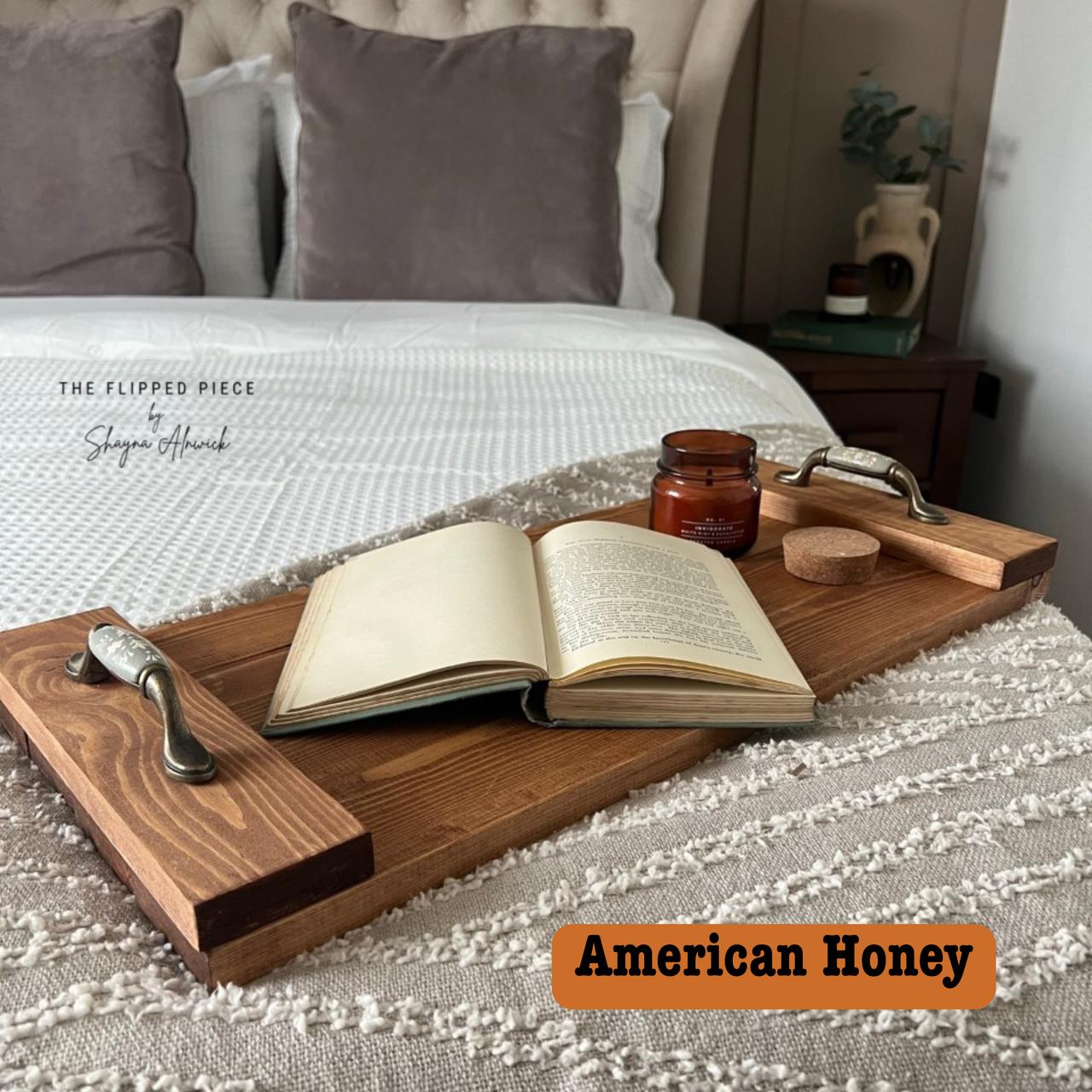 A cozy bedroom scene with an open book on a wooden tray positioned on a textured bedspread. A lit candle and a cork coaster are also on the tray. A bed with tufted pillows is in the background. Text reads "American Honey."