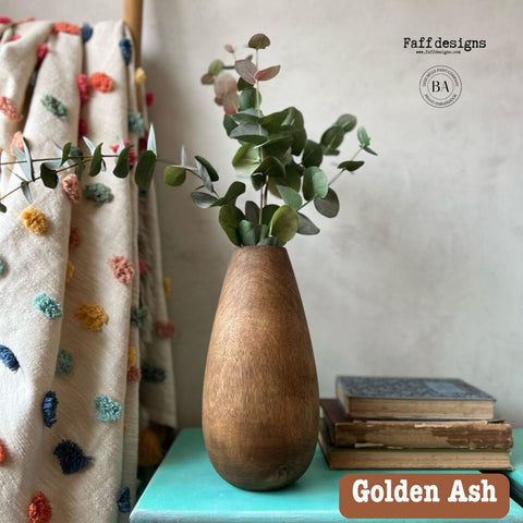 A wooden vase with eucalyptus branches is on a turquoise table next to stacked books. A colorful, textured curtain hangs beside it. The text reads "Faff designs" and "Golden Ash."