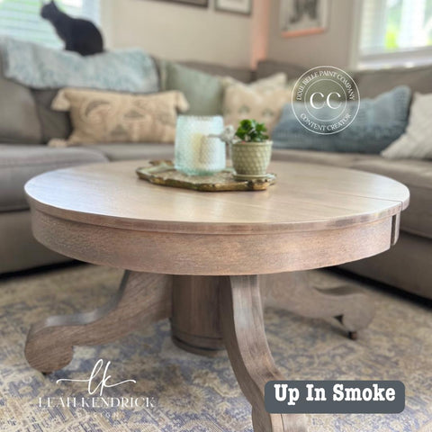 A wooden round coffee table on a patterned rug, holding a tray with a candle and small plant. A cat sits on the sofa in the background. Text reads "Up In Smoke," with logos for Leah Kendrick Design.